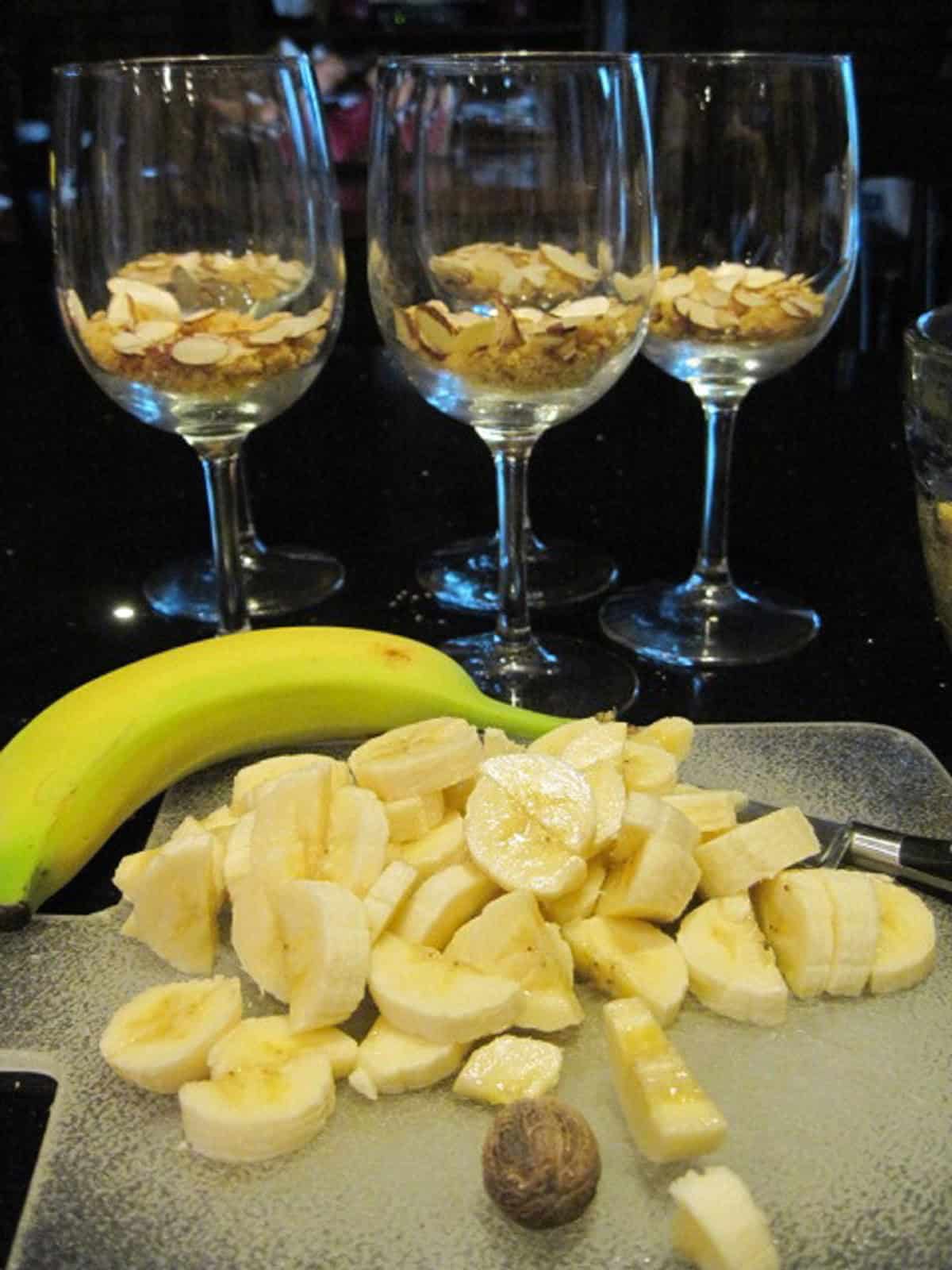 Three wine glasses in the background with sliced bananas in the foreground for making Banana Panna Cotta.