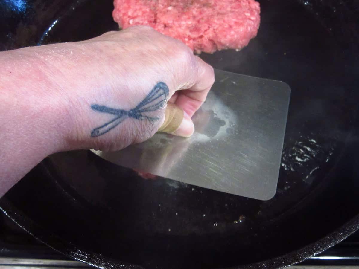 A hand pressing a burger with a spatula in a cast iron skillet.