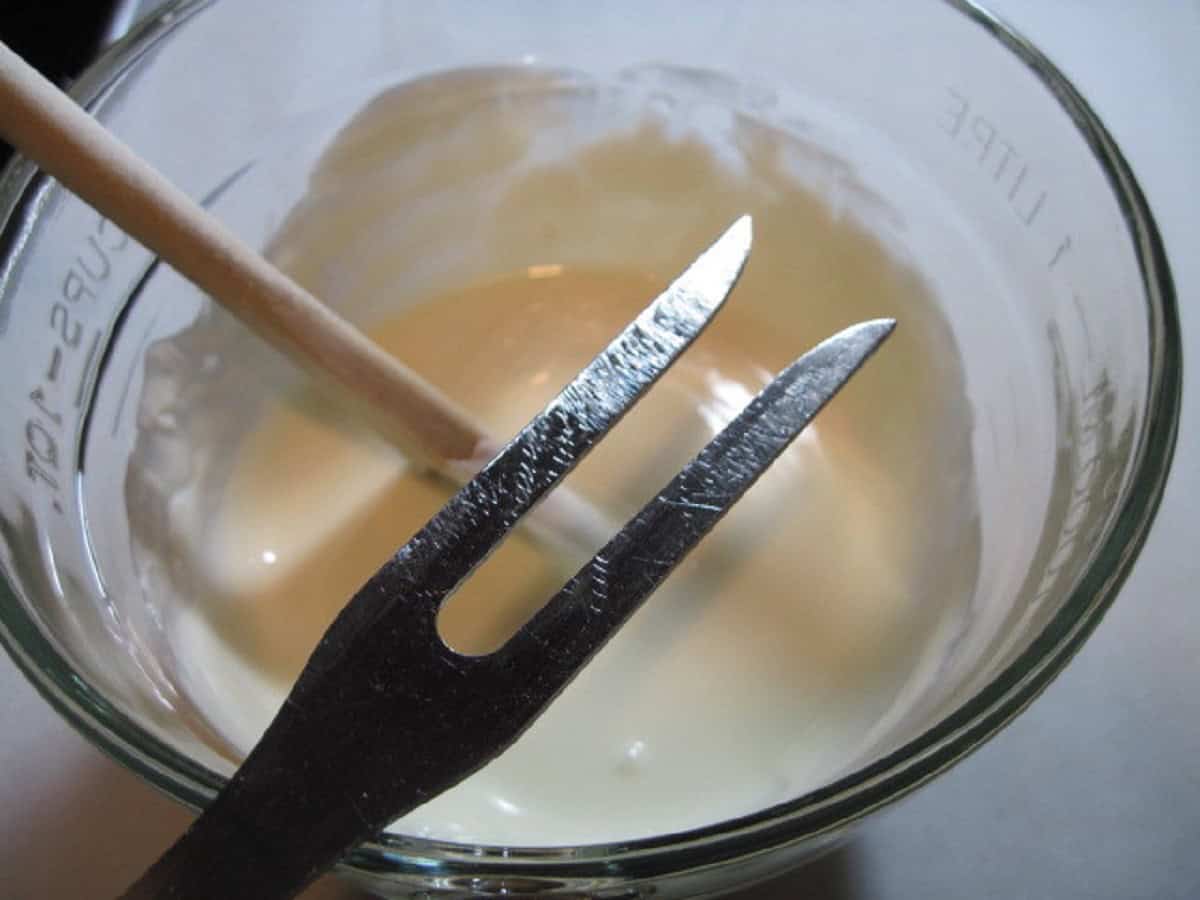 A meat fork being held over a bowl of melted white chocolate.