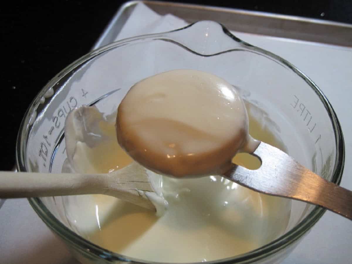 A cookie dipped in melted white chocolate over a glass bowl.