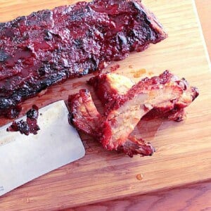 A wooden board with Slow Roasted Baby Back Ribs and a cleaver.