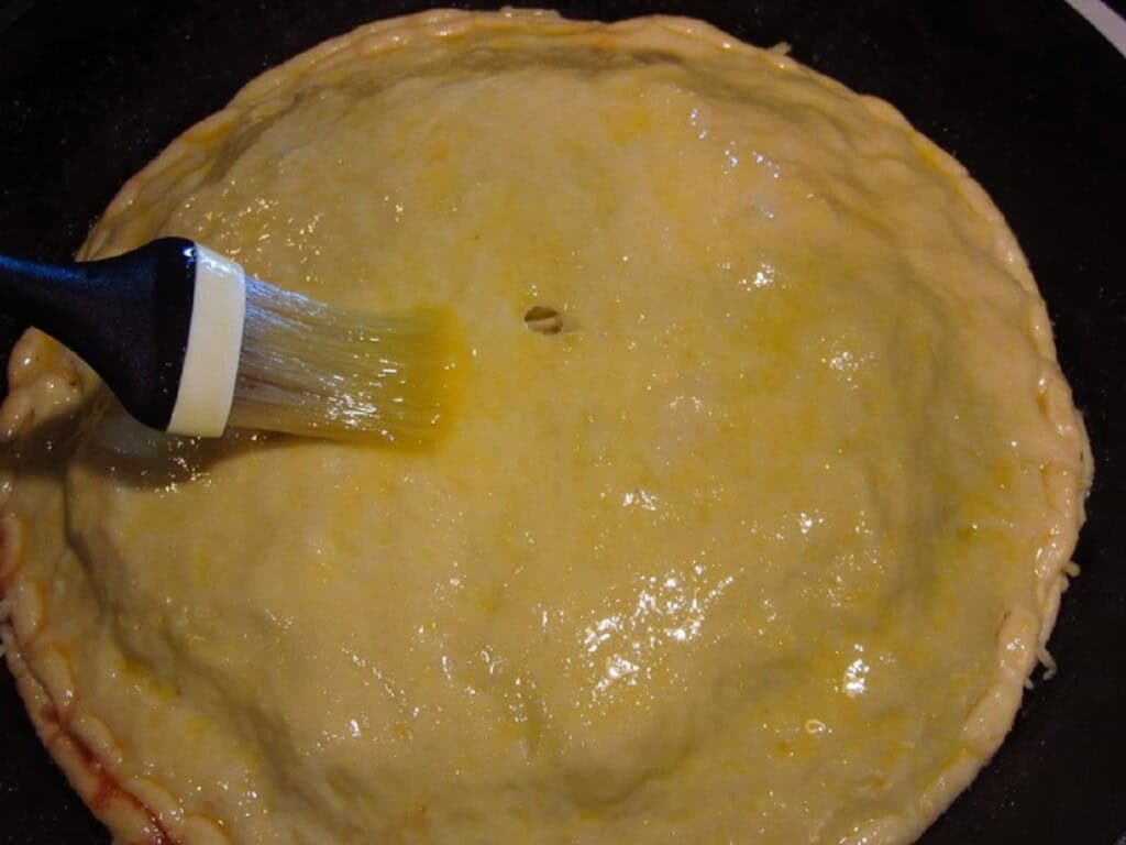A pastry brush brushing egg wash on the crust of a Hawaiian pizza.