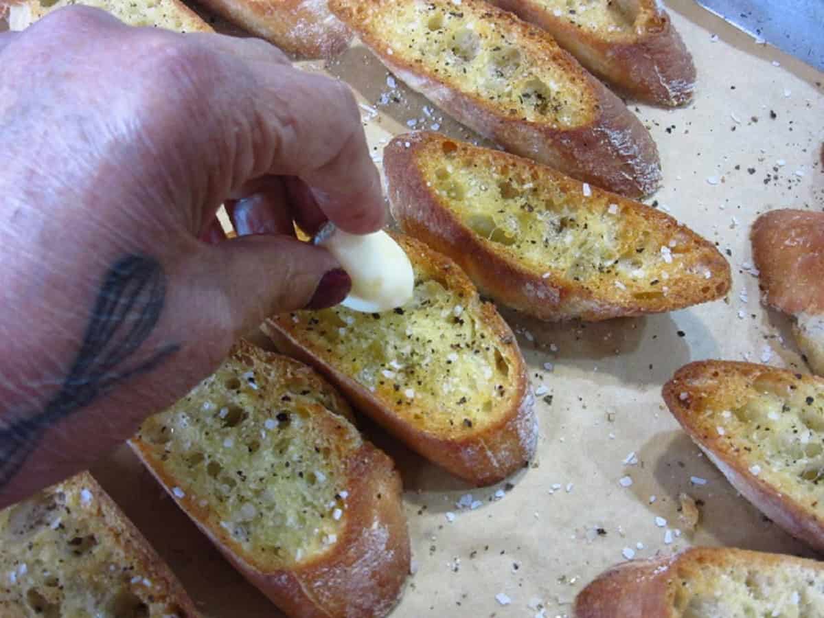 A garlic clove being rubbed onto a toasted crostini slice.