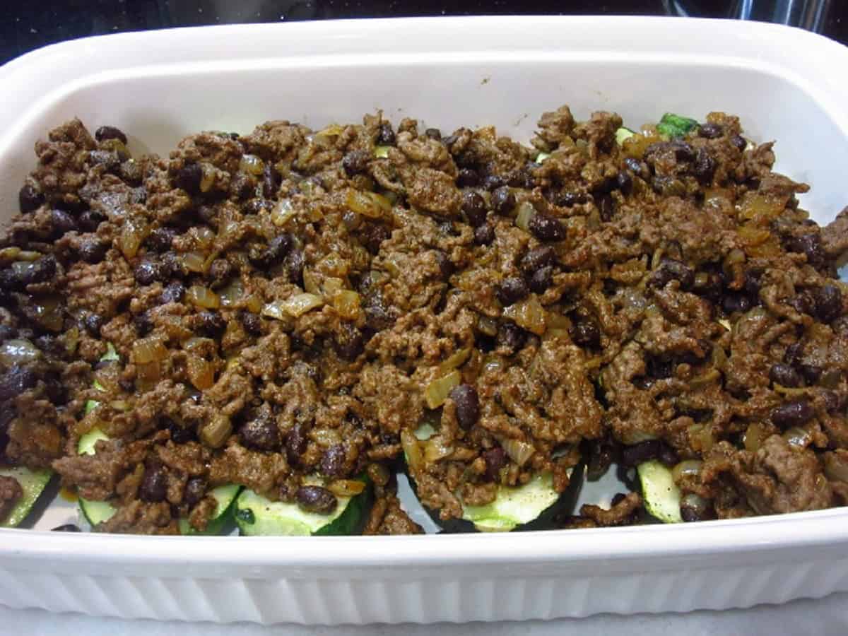 Cooked ground beef on top of zucchini boats in a white casserole dish.