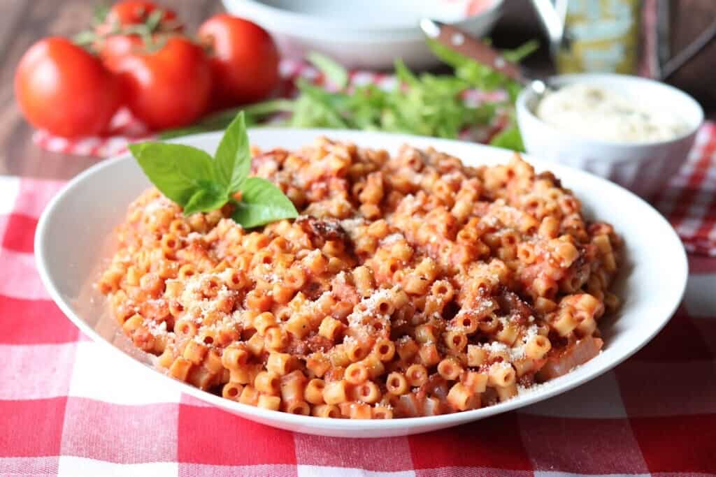 A red and white tablecloth with a white bowl filled with Tomato Bacon Pasta with Mascarpone Sauce.