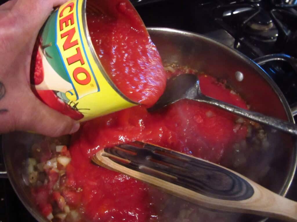 A can of crushed tomatoes being added to a skillet of bacon and onions.