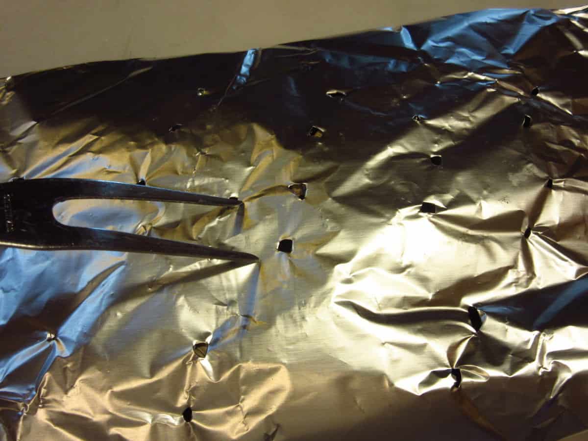 A meat fork poking holes into a packet of wood chips wrapped in foil to turn an outdoor grill into a smoker.