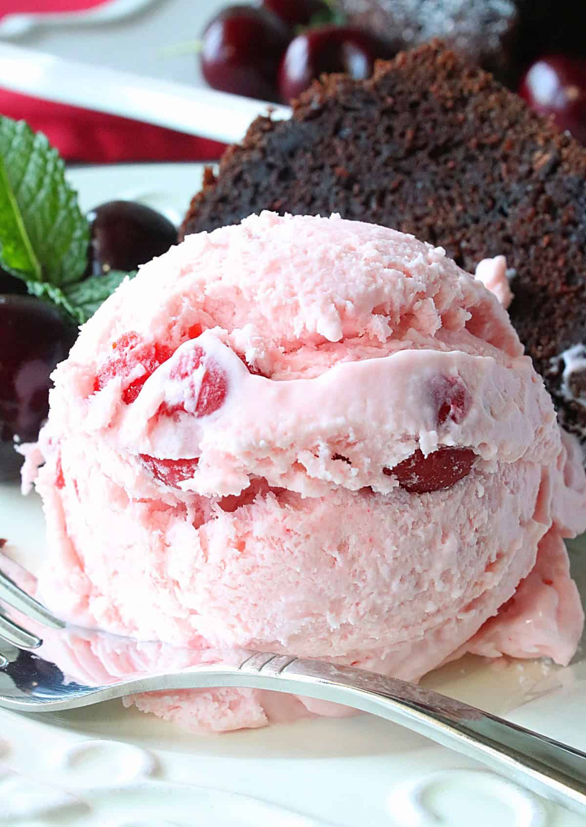 A round scoop of Cherry Amaretto Ice Cream on a white plate with chocolate cake in the background.
