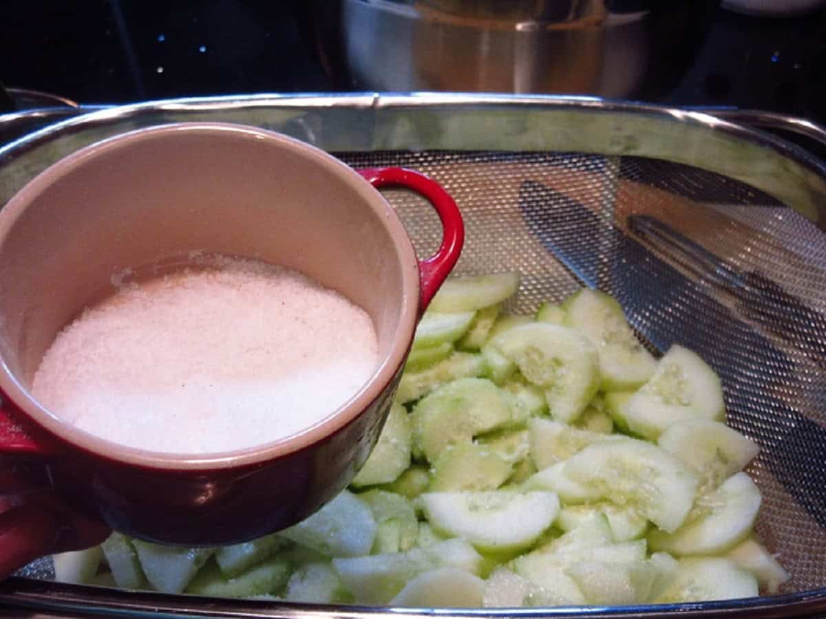 A container of salt over a strainer full of chopped cucumbers.