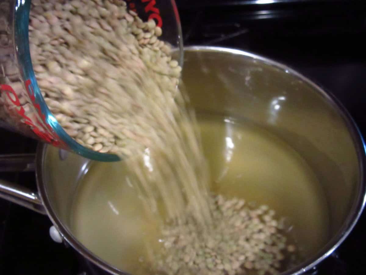 Brown lentils being poured into a stockpot with broth.