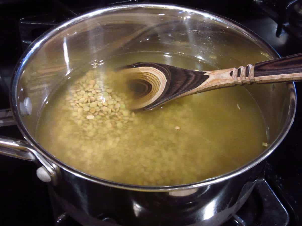 Brown lentils in a saucepan with stock and a wooden spoon.