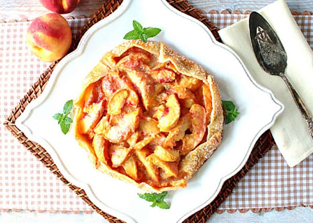 A scalloped white plate with a round peach crostata and fresh mint garnish.