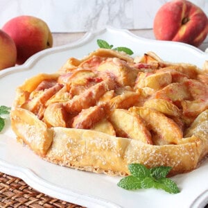 A pretty fresh peach crostata on a plate with fresh peaches in the background.