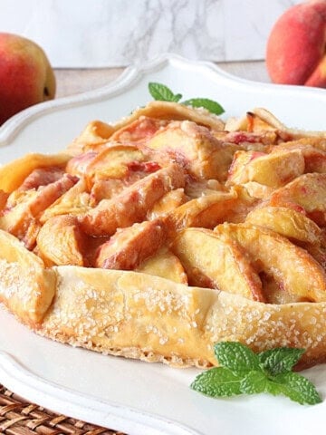A pretty fresh peach crostata on a plate with fresh peaches in the background.