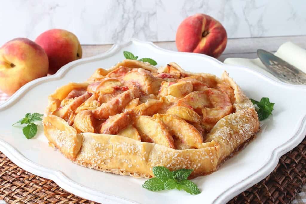 An entire round peach galette garnished with fresh mint leaves on a pretty white serving platter.