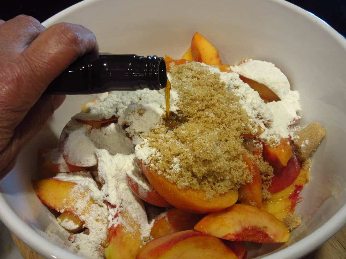Vanilla extract being poured into a bowl with fresh peaches and brown sugar.