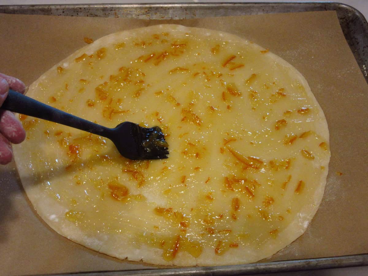 A pastry brush brushing out orange marmalade on a round pie crust.