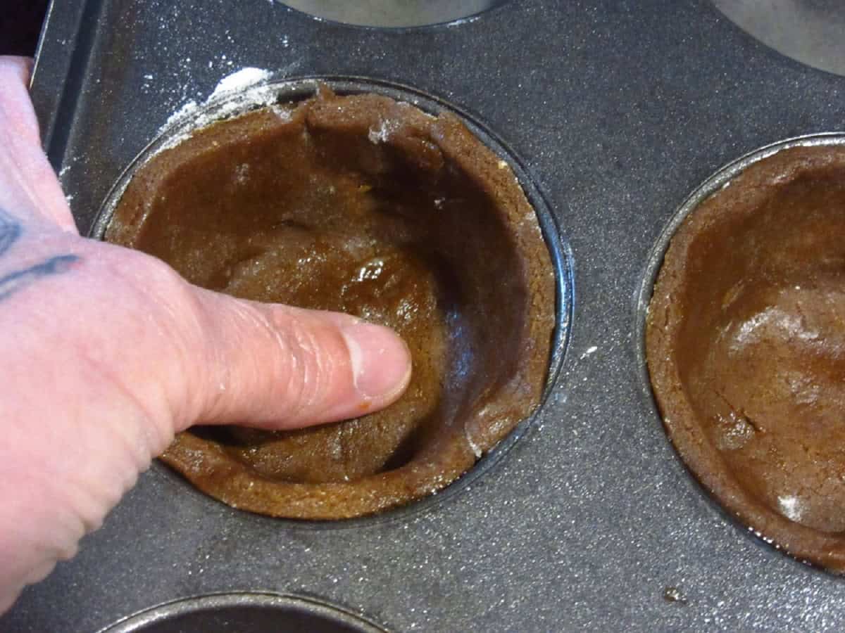 A thumb pressing gingerbread pie crust into a muffin tin.
