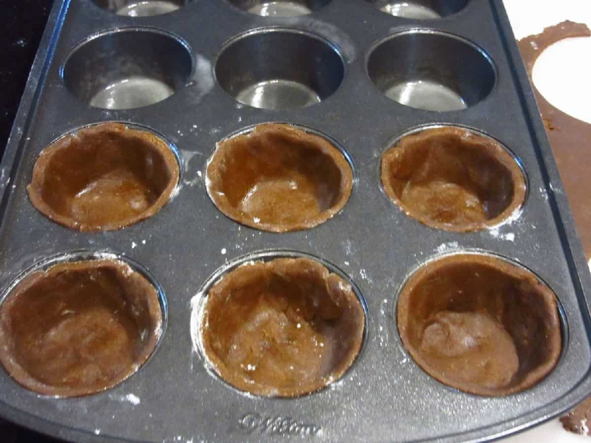 Six unbaked gingerbread pie crusts in a muffin tin.