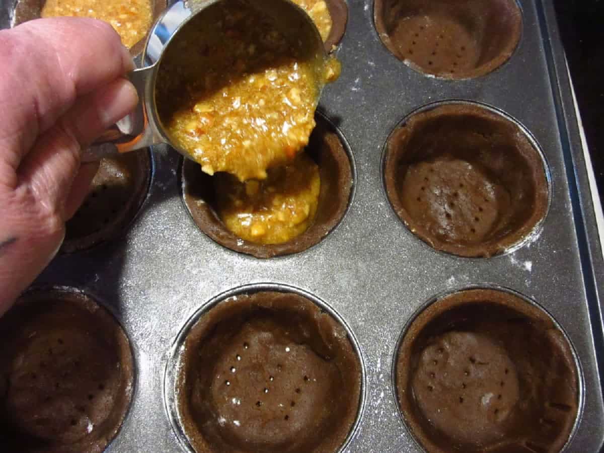 Pecan pie filling being poured into gingerbread pie tart shells.