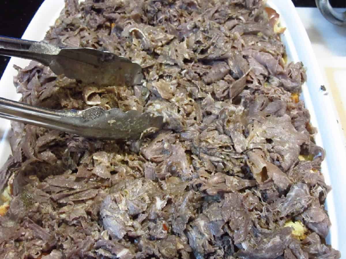 Chopped Italian beef in a casserole dish with tongs.