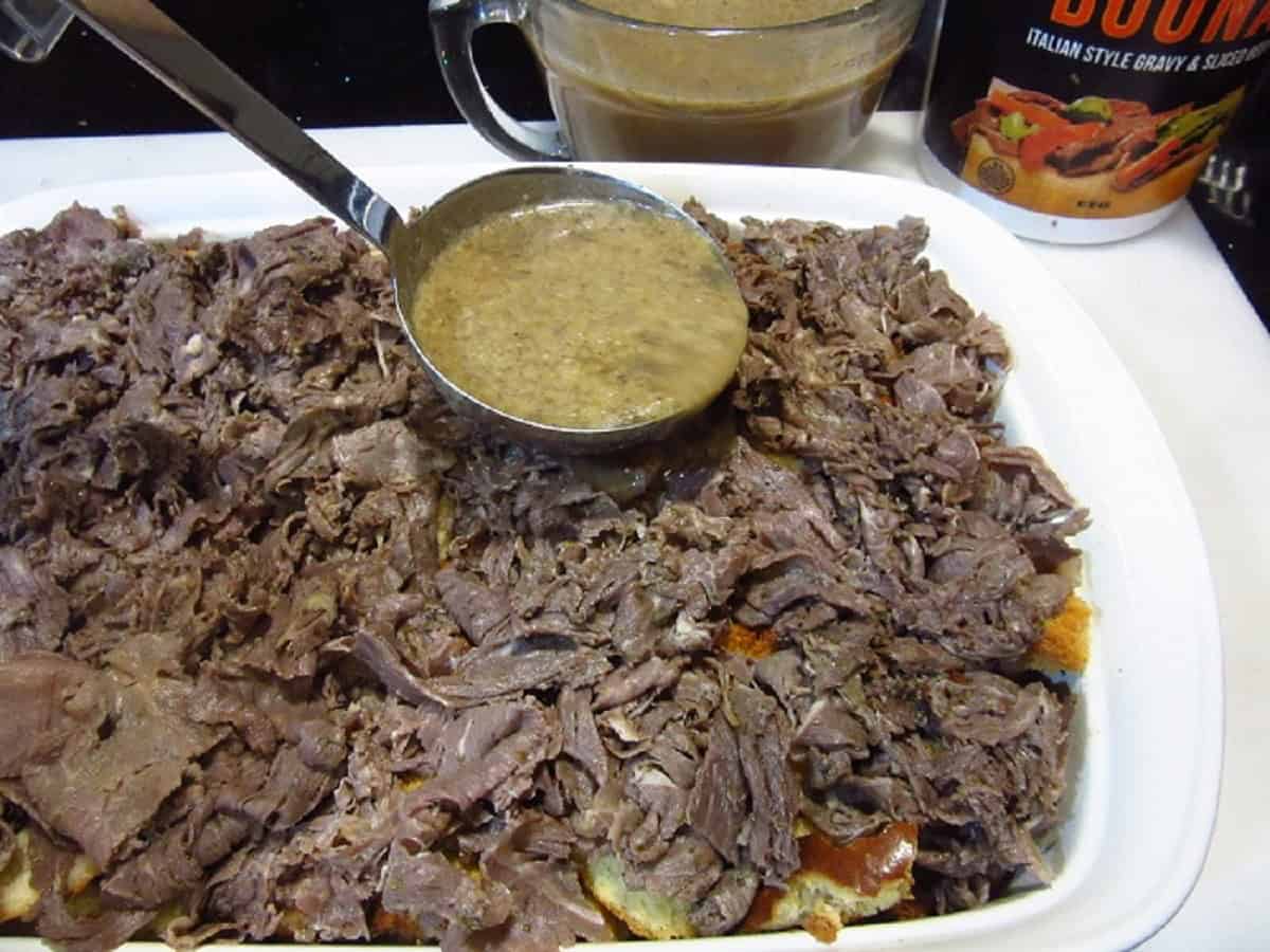 Gravy being spooned over Italian beef in a casserole dish.