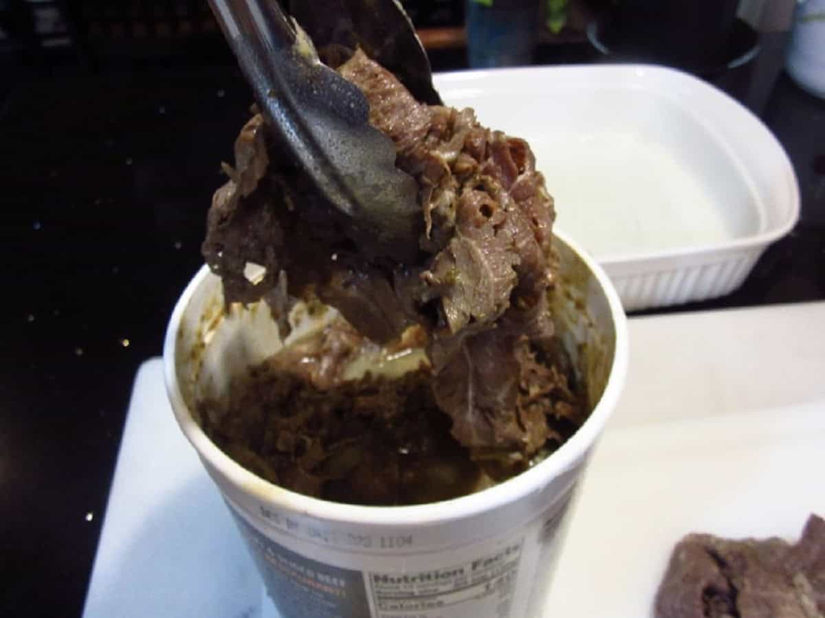 Tongs taking Italian beef out of a plastic tub.