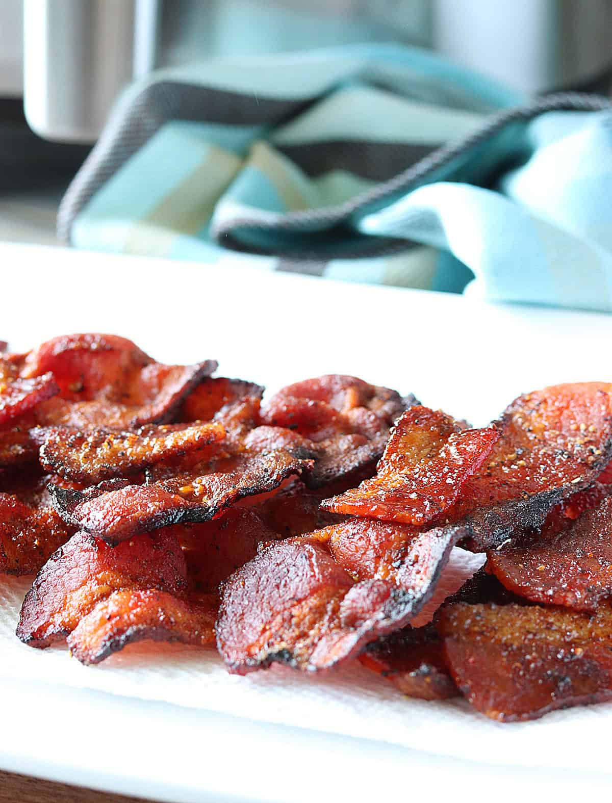 A white plate filled with air fryer bacon in the foreground and a blue napkin and air fryer in the background.