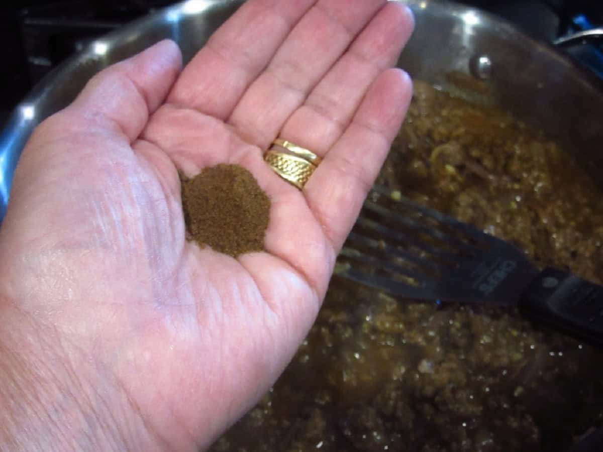 A hand holding a teaspoon of allspice over a large skillet.