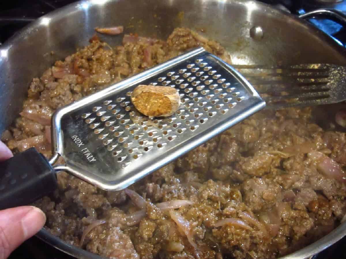 A pod of nutmeg being ground into a skillet of ground beef.