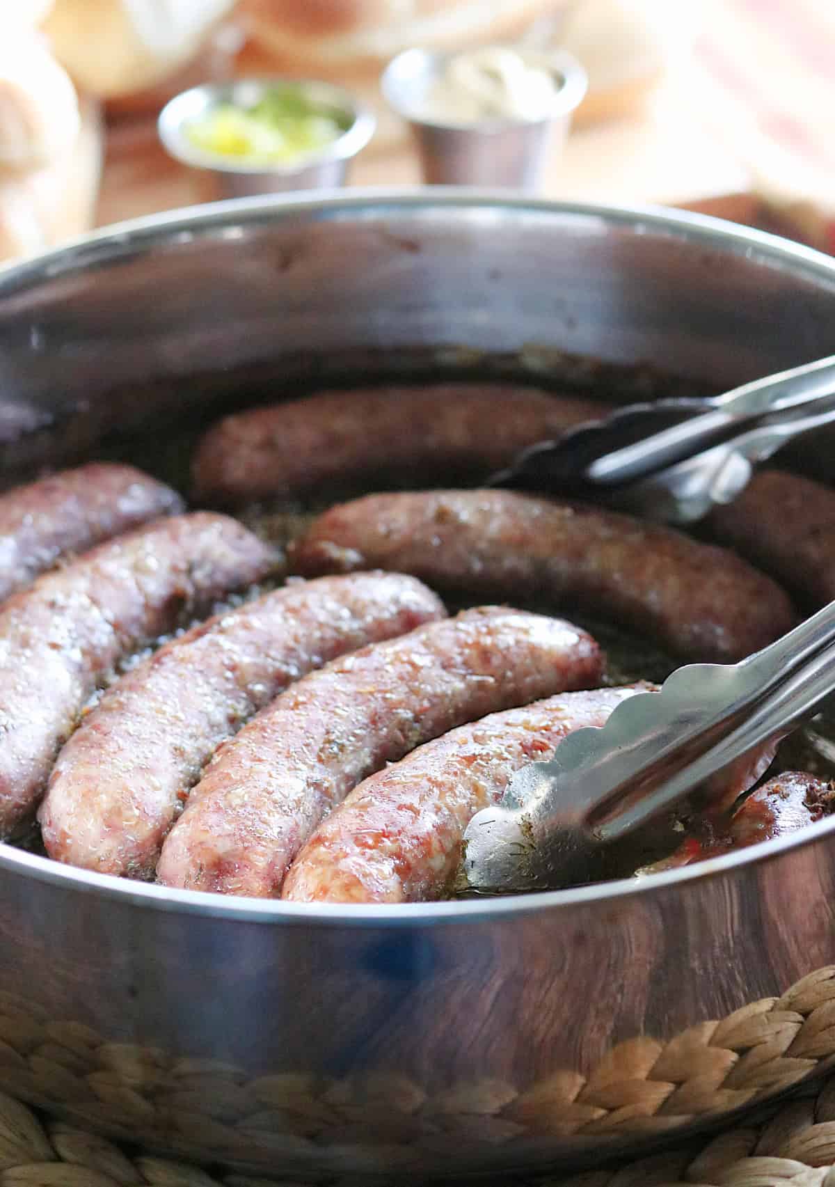 Cooked Bratwurst in a skillet with a pair of tongs.