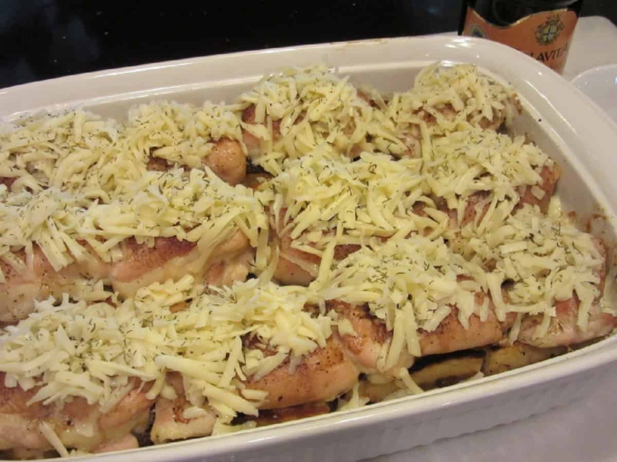 Shredded white cheese on top of baked chicken thighs in a white casserole dish.