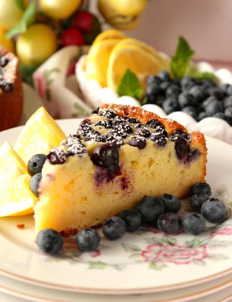 A slice of lemon ricotta cake on a pretty plate with fresh blueberries and lemon slices.