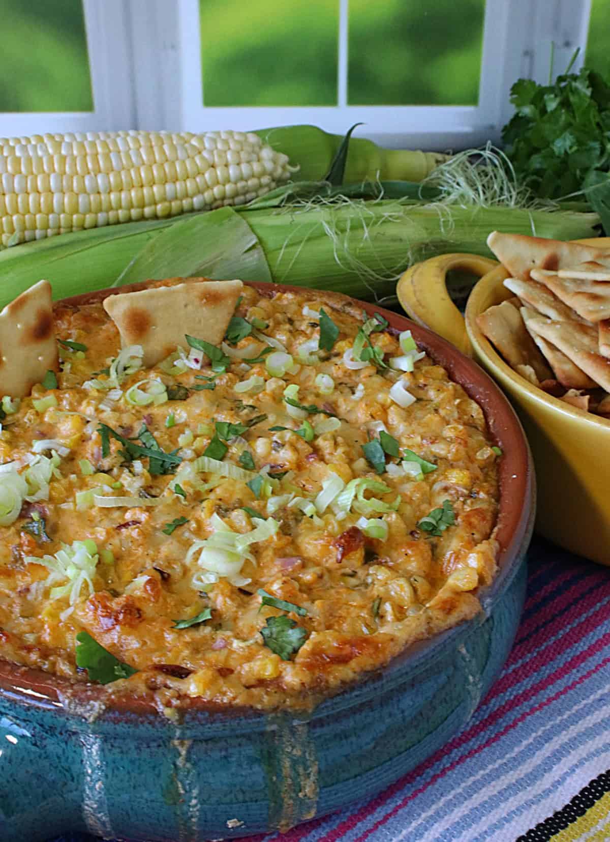 A blue crock filled with hot cheesy Mexican Street Corn Dip along with whole corn on the cob in the background.