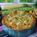 A blue Mexican crock filled with baked Cheesy Hot Mexican Street Corn Dip with some ears of corn in the background.