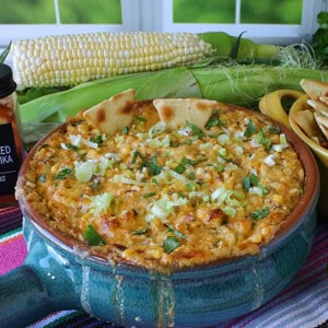 A blue Mexican crock filled with baked Cheesy Hot Mexican Street Corn Dip with some ears of corn in the background.