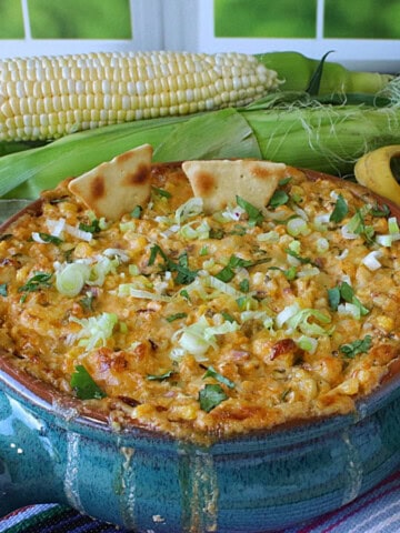 A blue Mexican crock filled with baked Cheesy Hot Mexican Street Corn Dip with some ears of corn in the background.