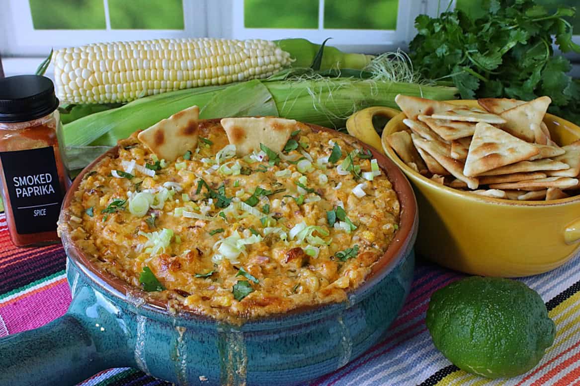 A blue crock of hot Mexican Street Corn Dip along with a bowl of pita chips and a lime.