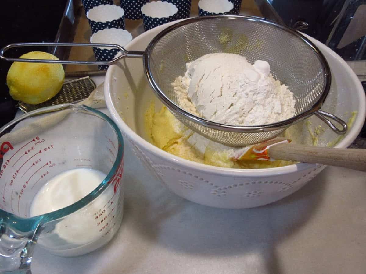 Flour in a strainer over a bowl of blueberry muffin batter.