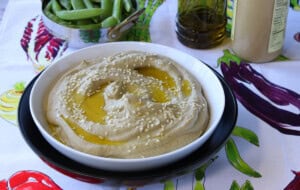 A colorful vegetable tablecloth with a bowl of Roasted Eggplant and Chickpea Hummus.