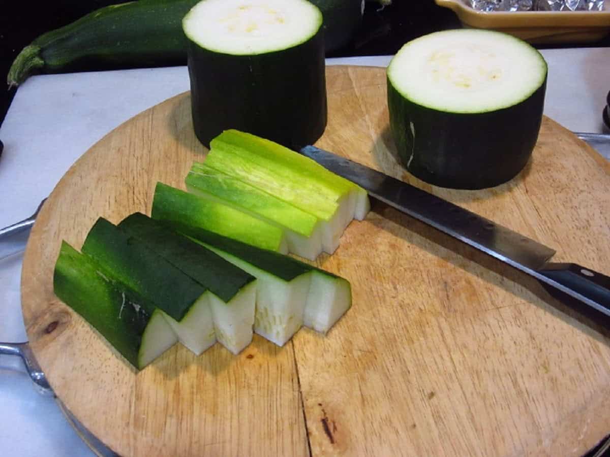 A zucchini being cut into chunks on a cutting board with a knife.