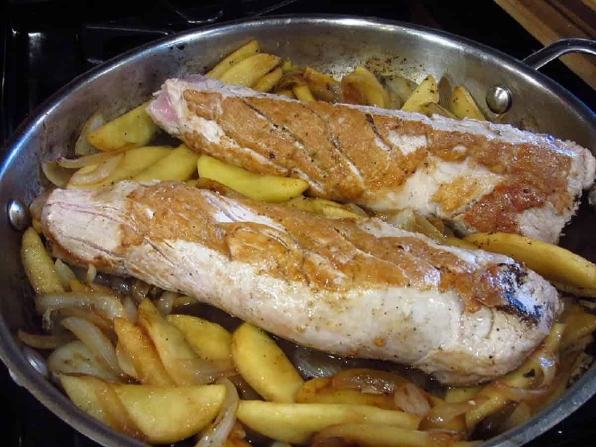 Two browned pork tenderloin in a skillet with apples and onions.