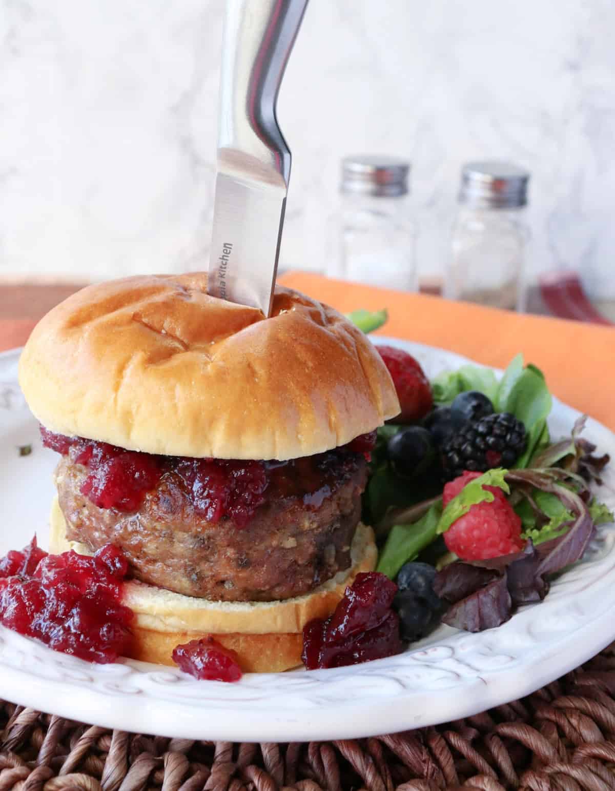 A knife stuck into a Stuffing Stuffed Turkey Burger on a white plate with a bun.