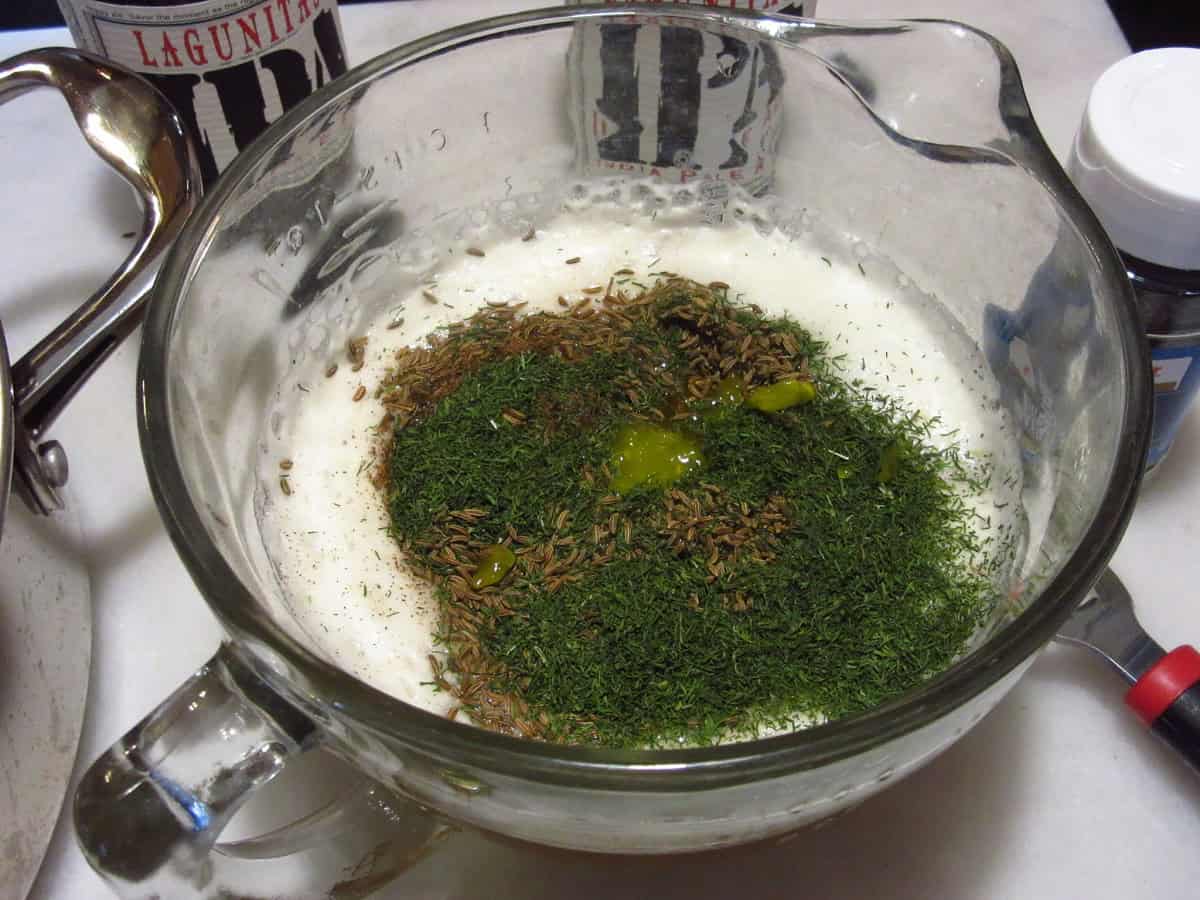 A glass measuring cup with caraway seed, dill, beer, and relish inside.