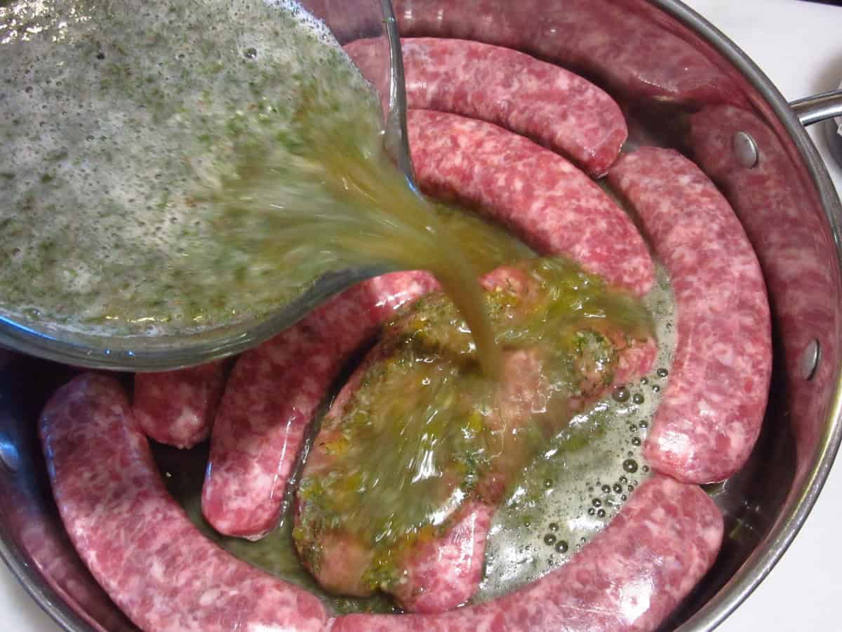 A braising liquid of beer and dill being poured over bratwurst in a skillet.