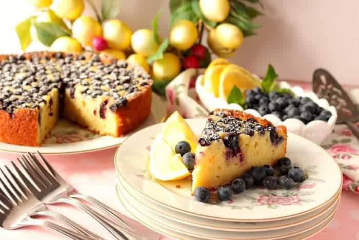 A slice of lemon ricotta cake on a plate with the whole cake in the background with lemons and blueberries as garnish.