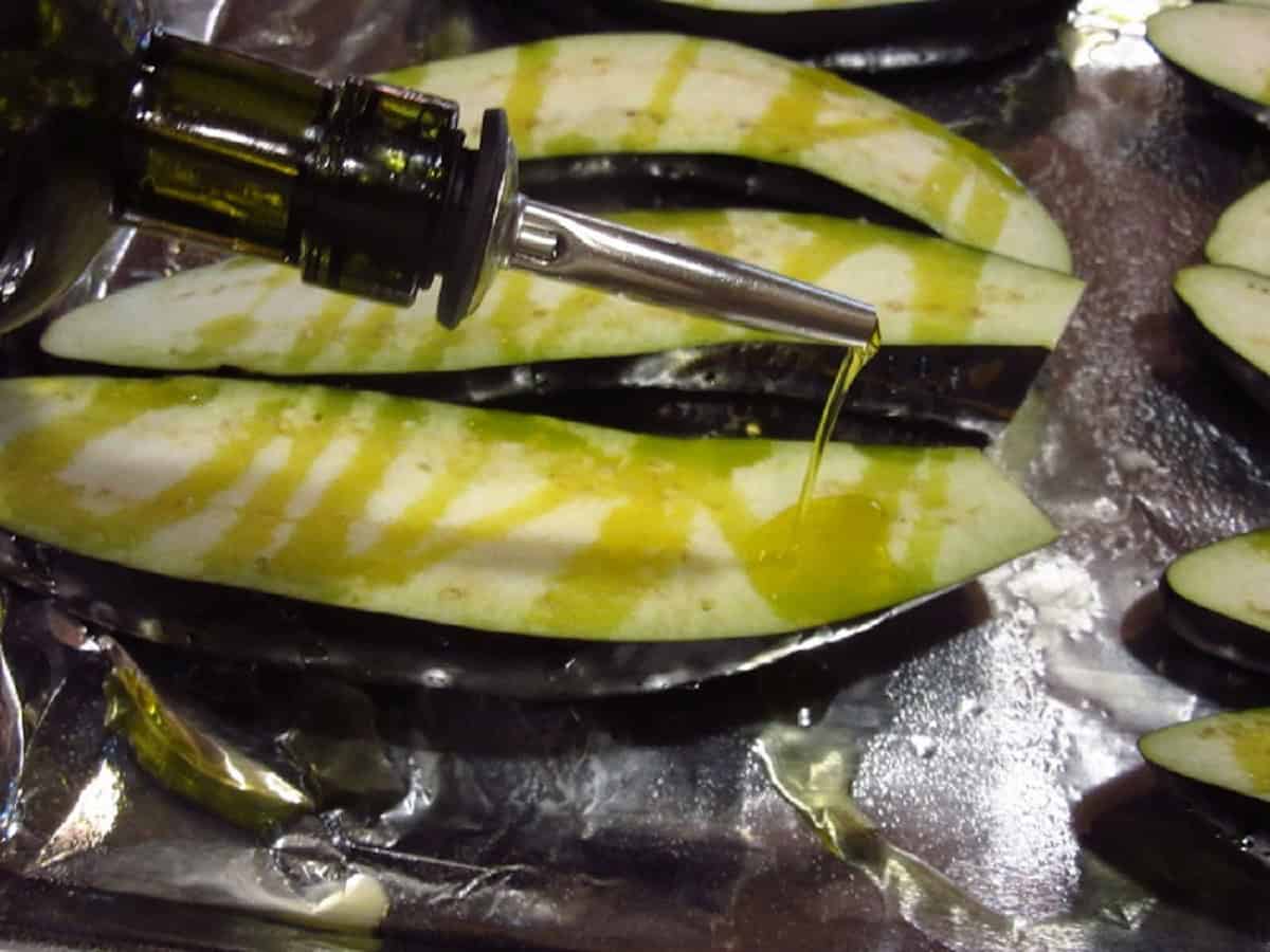 Olive oil being drizzled onto sliced eggplants on a baking sheet.