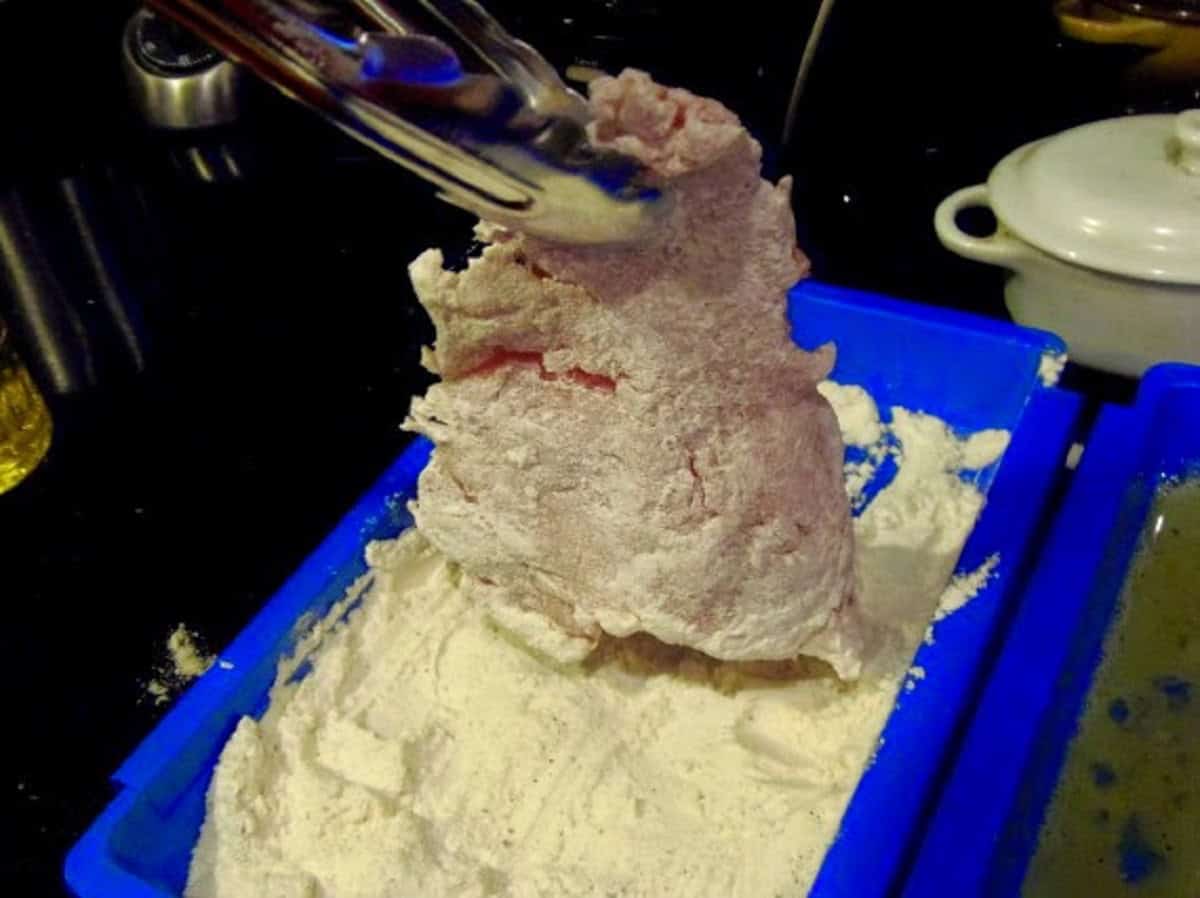 A pair of tongs holding up a floured pork cutlet for making schnitzel.