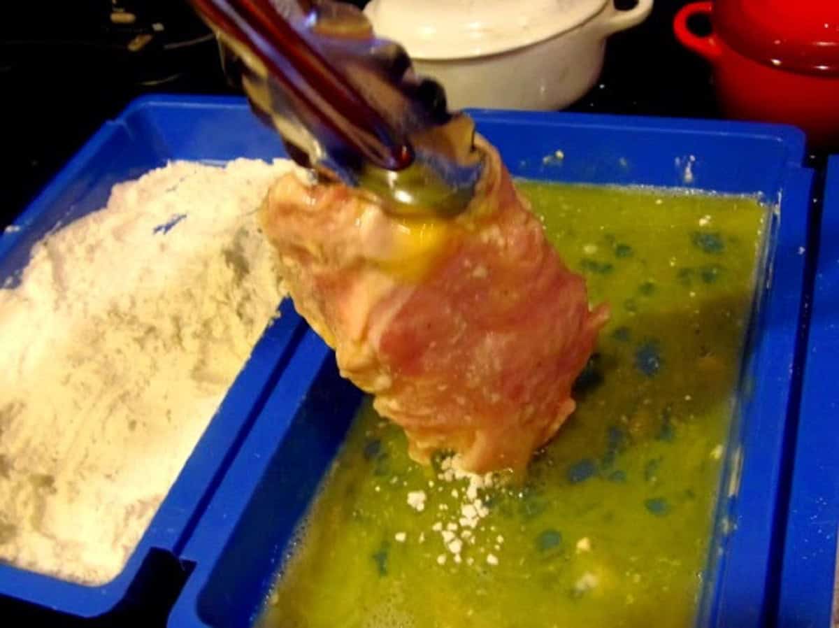 A pair of tongs holding up an egg wash pork cutlet for making pork schnitzel.