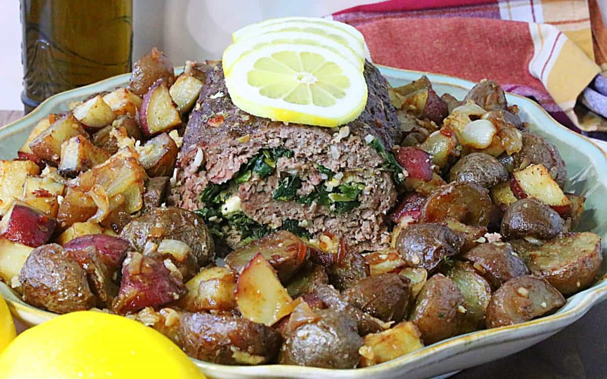 A Rolled Greek Meatloaf on a serving platter surrounded by golden brown potatoes.